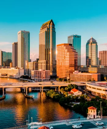 Aerial view of a vibrant cityscape at sunset with a mix of modern high-rise buildings and office towers reflecting golden sunlight. The image also features a calm river with several bridges and lush greenery. Vehicles are visible on the roads and bridges. - Hausion