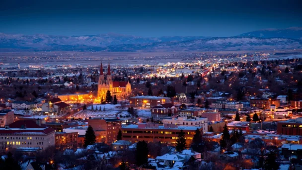 A panoramic view of a city at dusk, with buildings and streets lit up. A prominent illuminated cathedral stands out in the middle of the scene. Snow-covered rooftops and distant mountain ranges are visible under a deep blue sky. - Hausion