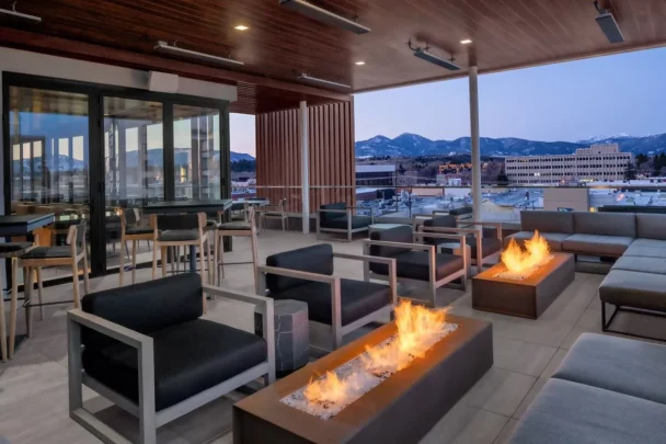 A modern rooftop lounge with contemporary outdoor furniture centered around rectangular fire pits. The space features tall tables and chairs along with cushioned seating. The backdrop includes a view of a cityscape and distant mountains under a dusk sky. - Hausion