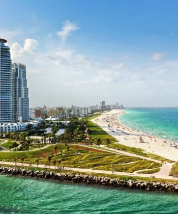 Aerial view of a coastal city featuring a curving beach with green parkland. A tall building with a red top dominates the shoreline, while smaller buildings stretch inland. The beach is bustling with people, and turquoise waters extend to the horizon under a blue sky. - Hausion