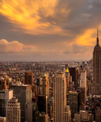 A cityscape of New York City during sunset. The image showcases numerous high-rise buildings, including the iconic Empire State Building in the foreground. The sky is painted with golden clouds, reflecting the warm hues of the setting sun. - Hausion