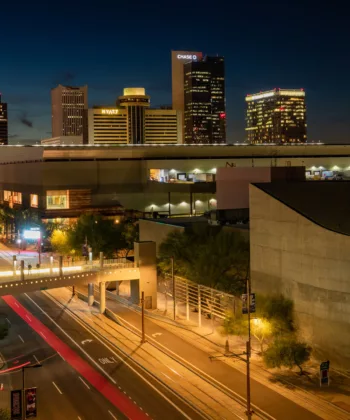 Nighttime cityscape of downtown Phoenix, Arizona, featuring illuminated skyscrapers, light trails from moving vehicles, and a prominent building with "Arizona Science Center" signage. Streetlights and pedestrian bridges add architectural interest to the scene. - Hausion