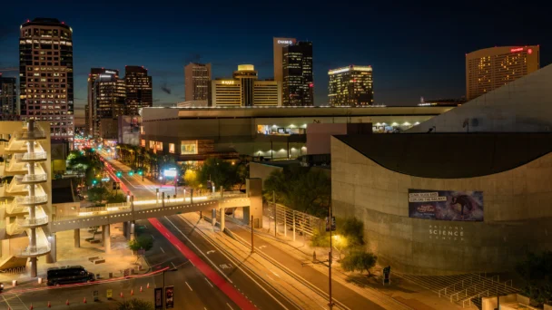 Nighttime cityscape of downtown Phoenix, Arizona, featuring illuminated skyscrapers, light trails from moving vehicles, and a prominent building with "Arizona Science Center" signage. Streetlights and pedestrian bridges add architectural interest to the scene. - Hausion