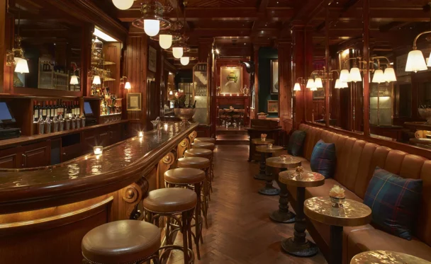 A dimly lit, elegant bar with dark wood paneling and plush leather seating. The curved bar features ornate details with barstools lined up. Tables and couches to the right enhance the cozy atmosphere, illuminated by soft lights hanging from the ceiling. - Hausion