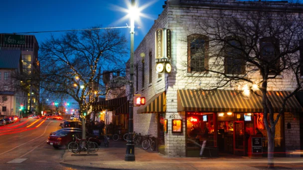 A busy street corner at dusk in a downtown area. A brightly lit restaurant with awnings and signs occupies the corner, displaying "Open" signs. Bikes are parked outside, and string lights are wrapped around leafless trees. Traffic lights and light trails from cars are visible. - Hausion