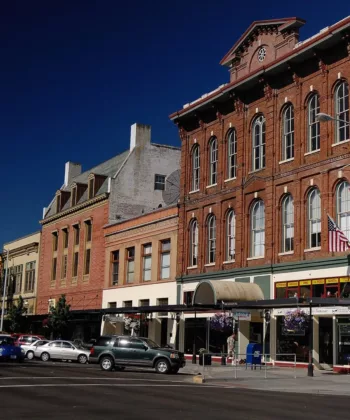 A wide street view of a downtown area with historic brick buildings under a clear blue sky. A tall white building stands in the background, surrounded by shorter structures. Cars are parked along the street, and small shops are visible at street level. - Hausion