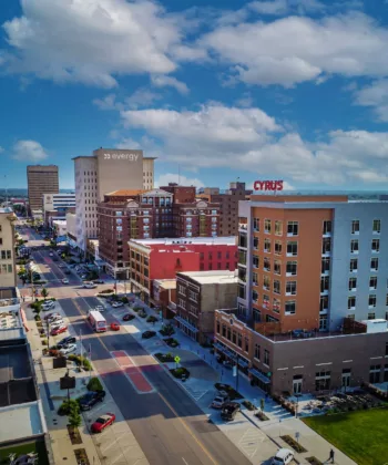 A vibrant urban cityscape shows a street lined with mid-rise buildings, including the prominently labeled Evergy and Cyrus buildings. Cars and people are visible on the street, and the sky is bright with fluffy clouds. - Hausion