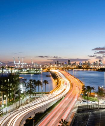 A vibrant cityscape at dusk featuring a busy highway with streaks of headlights and taillights. The road is flanked by towering buildings, and a body of water with cranes and a distant city skyline is visible against a colorful twilight sky. - Hausion