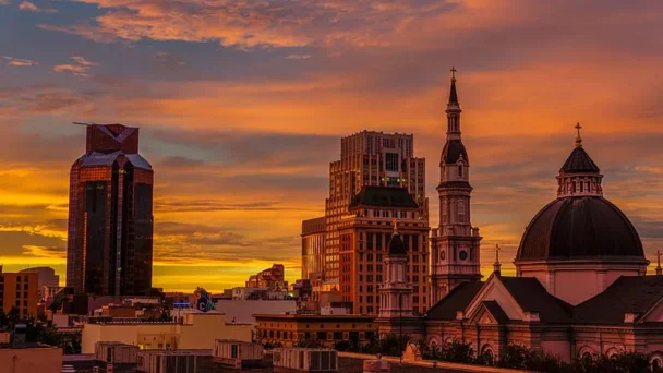 A city skyline at sunset featuring a mix of modern and historic architecture. Prominent buildings include a tall, glass skyscraper with a unique design, a church with a domed roof and a spire, and other varied structures beneath a vibrant, orange sky. - Hausion