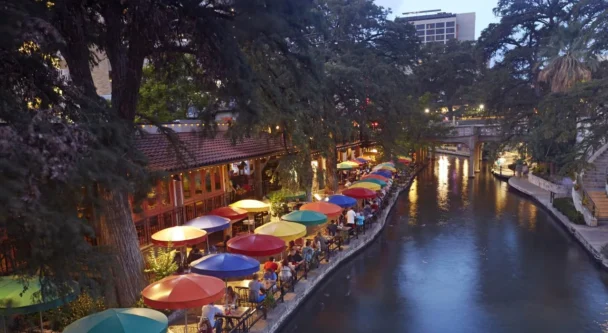 A lively riverside scene at dusk, featuring colorful umbrellas covering outdoor tables along a restaurant-lined walkway. The river reflects the evening lights, and lush trees frame the area. People are dining and strolling along the scenic riverbank. - Hausion