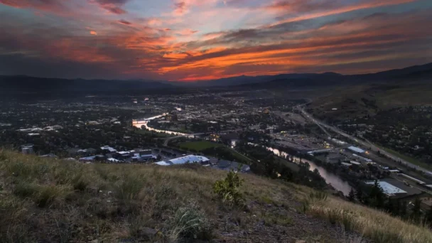 A cityscape at dusk with a vibrant orange and purple sunset. The city is nestled in a valley, surrounded by hills and mountains. A river cuts through the city, reflecting the colors of the sky. Grassy terrain is visible in the foreground. - Hausion