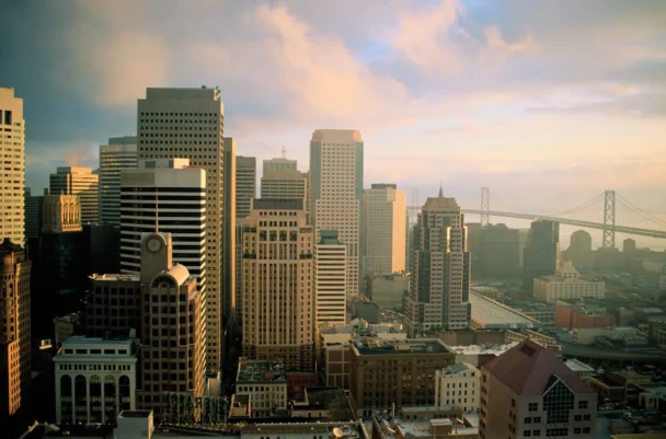 An aerial view of a cityscape at sunrise, featuring tall skyscrapers with varying architectural styles. The sky is partly cloudy, and a large suspension bridge can be seen in the background, spanning across a body of water. - Hausion