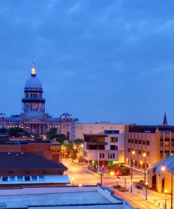 A cityscape at dusk showcases well-lit buildings, streets, and a prominent domed structure in the background. The sky is a deep blue with scattered clouds, adding to the serene evening ambiance. - Hausion