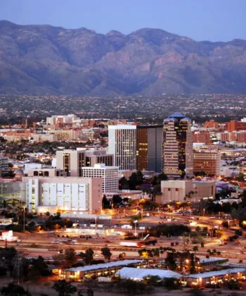 Aerial view of Tucson, Arizona at dusk, showcasing a mix of modern and historic buildings. The city's skyline is backed by the rugged mountain range under a transitioning sky from day to night, with lights starting to illuminate the area. - Hausion