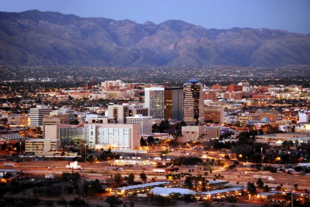 Aerial view of Tucson, Arizona at dusk, showcasing a mix of modern and historic buildings. The city's skyline is backed by the rugged mountain range under a transitioning sky from day to night, with lights starting to illuminate the area. - Hausion