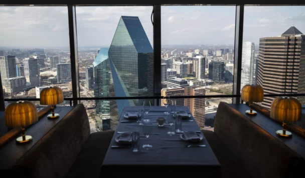 A restaurant table set for four, located by a large window with a panoramic view of a city's skyline. Prominent buildings, including a distinctive green glass pyramid-like structure, are visible. The table has yellow lamp shades and is elegantly arranged. - Hausion