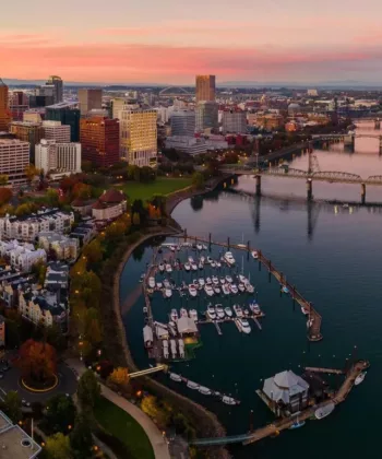 Aerial view of a cityscape at sunset featuring a river with multiple bridges and a marina with boats docked. Modern and historic buildings, green spaces, and tree-lined streets are visible, with a mountainous backdrop and a sky filled with hues of pink and orange. - Hausion