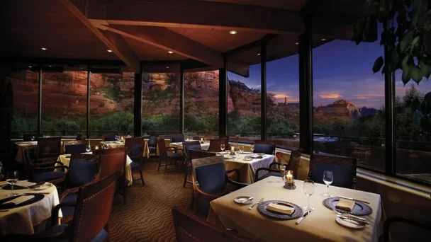 A cozy restaurant with elegantly set tables, each adorned with candles, plates, and glasses. The floor-to-ceiling windows offer stunning views of a rocky desert landscape at dusk, with the sky painted in deep blue and purple hues. - Hausion