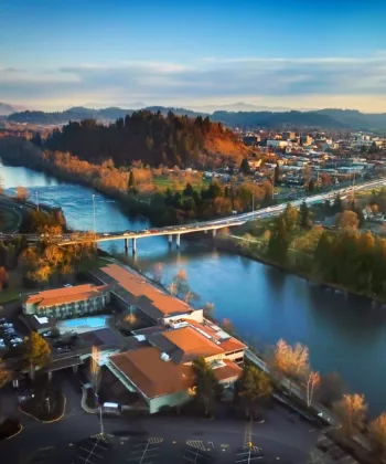 Aerial view of a picturesque town with a prominent bridge spanning a river. Surrounding areas include buildings, roads, and green spaces, with hills and a partly cloudy sky in the background. The scene is bathed in warm, late afternoon sunlight. - Hausion