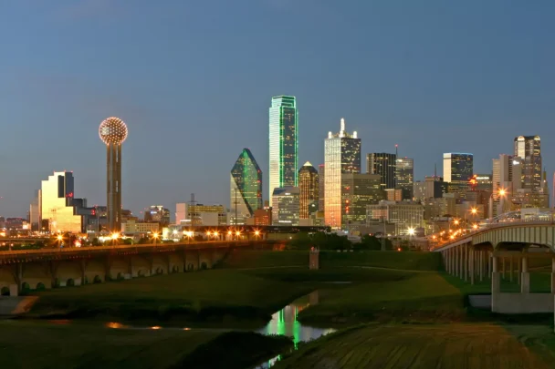 A nighttime view of the Dallas skyline, showing illuminated skyscrapers including the green-lit Bank of America Plaza and Reunion Tower with its spherical lights. The city lights reflect on the river and the sky subtly transitions from dusk to night. - Hausion