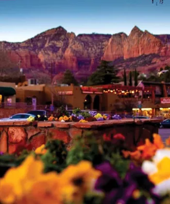 A vibrant street scene in Sedona, Arizona, featuring colorful flowers in the foreground, shops, restaurants, and parked cars in the midground, and a backdrop of stunning red rock formations under a blue sky at dusk. - Hausion