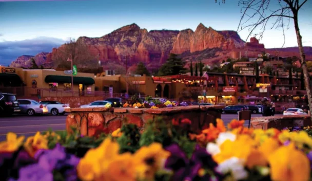 A vibrant street scene in Sedona, Arizona, featuring colorful flowers in the foreground, shops, restaurants, and parked cars in the midground, and a backdrop of stunning red rock formations under a blue sky at dusk. - Hausion