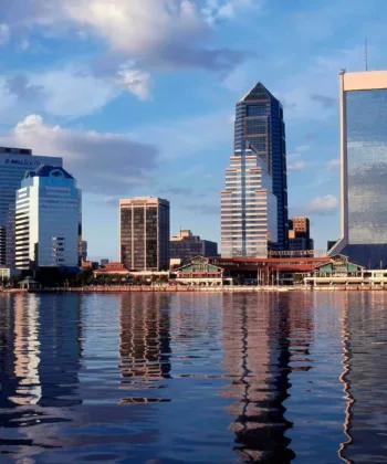 A city skyline along the water, featuring several modern skyscrapers and buildings with cloud reflections in the water below. The sky is partly cloudy, adding to the reflective effect on the calm water surface. - Hausion