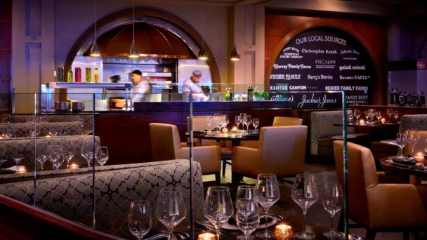 A dimly lit, elegant restaurant dining area with upholstered chairs and tables set with wine glasses and candles. A glass partition separates the diners from an open kitchen where chefs are at work. A large chalkboard lists local food sources in the background. - Hausion