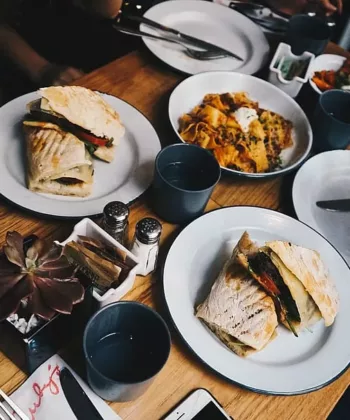 A table set for a meal with four plates containing different dishes, including sandwiches and a pasta dish. There are drinks in blue cups, a takeout coffee cup, and condiments in the center. Cutlery and a small succulent plant are also visible on the table. - Hausion