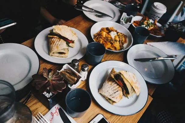 A table set for a meal with four plates containing different dishes, including sandwiches and a pasta dish. There are drinks in blue cups, a takeout coffee cup, and condiments in the center. Cutlery and a small succulent plant are also visible on the table. - Hausion