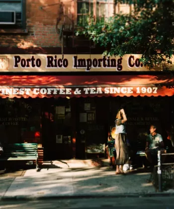 A small coffee shop called Porto Rico Importing Co. features a red awning with gold lettering reading "Finest Coffee & Tea Since 1907." People are sitting outside; one person stands near the entrance. Trees and shadows add a cozy, shaded ambiance. - Hausion