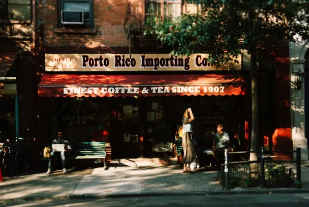 A small coffee shop called Porto Rico Importing Co. features a red awning with gold lettering reading "Finest Coffee & Tea Since 1907." People are sitting outside; one person stands near the entrance. Trees and shadows add a cozy, shaded ambiance. - Hausion