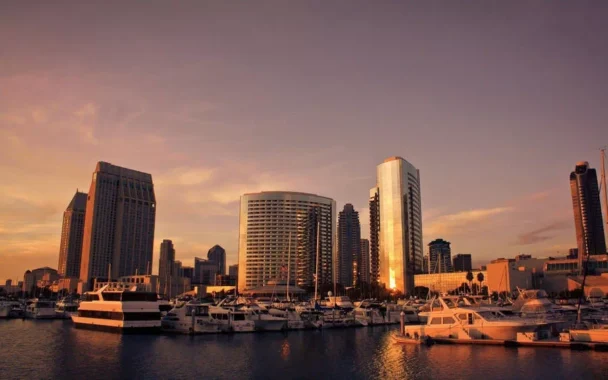 Sunset view of a city marina with several docked boats. The scene includes tall, modern buildings reflecting the golden light of the setting sun, with a clear sky and a peaceful body of water in the foreground. - Hausion