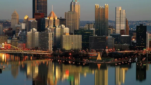 A vibrant cityscape at dusk featuring a skyline with various tall buildings, some illuminated. A river in the foreground reflects the city lights, and a brightly lit triangular structure stands near the water. Bridges and streets are visible amid the city lights. - Hausion