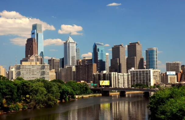 Philadelphia city skyline viewed from across the Schuylkill River, featuring a mix of modern and classic skyscrapers under a clear blue sky. A bridge spans the river, while lush green trees border the water, reflecting the cityscape. - Hausion