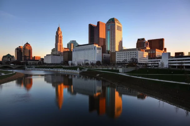 A city skyline at sunset is reflected in a calm river. Tall buildings with a mix of modern and historic architecture are illuminated by the golden light. Green spaces and walkways line the riverbank, enhancing the urban scenery. - Hausion