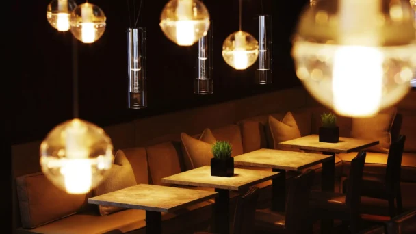 A dimly lit, modern cafe features a row of wooden tables with black chairs and beige cushions against a dark wall. Bright spherical pendant lights hang from the ceiling, casting a warm glow over the space. Small plants in black pots adorn each table. - Hausion