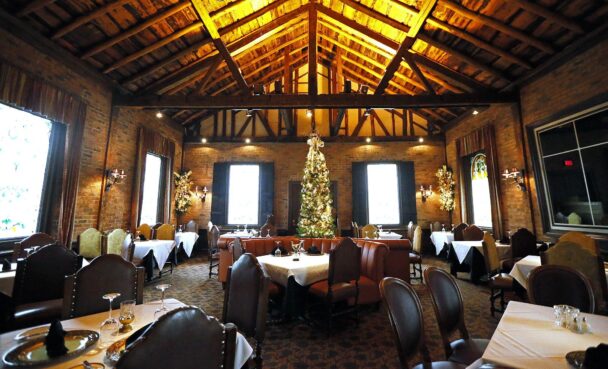 A cozy, rustic restaurant interior with brick walls and a high wooden-beam ceiling. The room is decorated for the holiday season with a large, beautifully adorned Christmas tree at the center. Tables are set with white tablecloths and elegant place settings. - Hausion