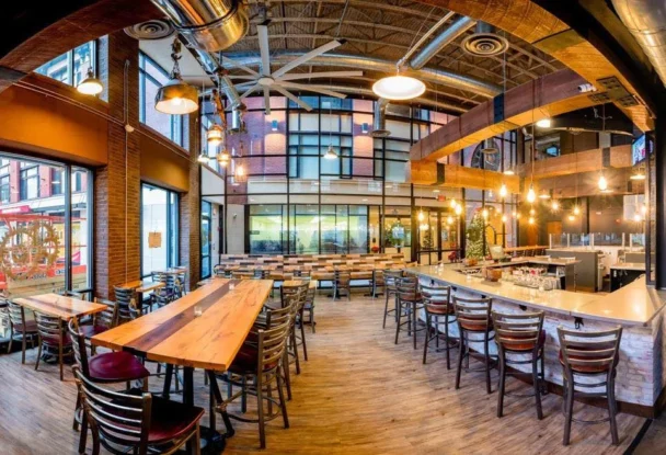 A well-lit, modern restaurant interior featuring wooden tables and chairs. The area includes a long bar on the right with high stools, exposed ductwork, and large windows letting in natural light. The decor blends industrial and rustic elements, creating a cozy atmosphere. - Hausion