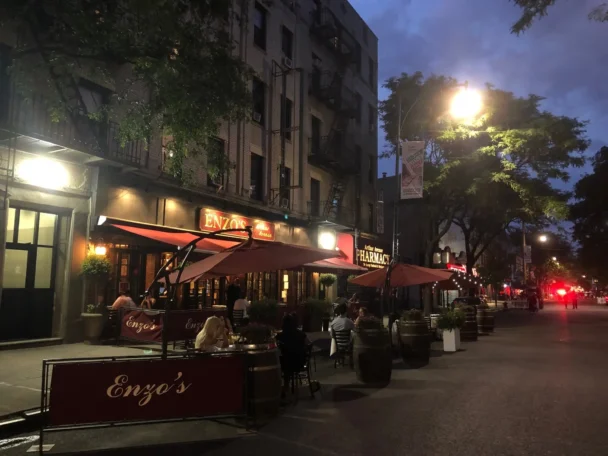 Street scene at dusk with an outdoor dining area in front of a restaurant named "Enzo's." Several people are seated under large red umbrellas. The surroundings include adjacent buildings, trees, and streetlights illuminating the area. - Hausion