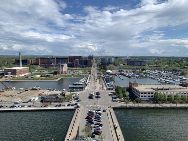 Aerial view of a harbor town showing a wide road stretching into the distance, flanked by buildings and parking spaces. Boats are docked along the water, and the sky is partly cloudy. Green spaces and trees are visible amidst the urban landscape. - Hausion