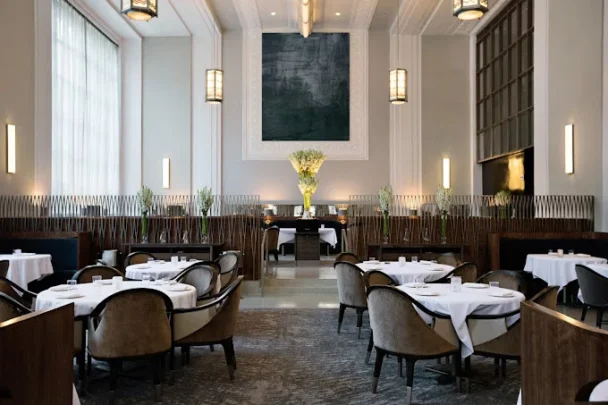 A spacious, elegant restaurant interior with high ceilings, large windows, and modern chandeliers. Round tables with white tablecloths and plush, grey chairs are neatly arranged. A large, dark artwork and numerous flower arrangements add sophistication to the setting. - Hausion