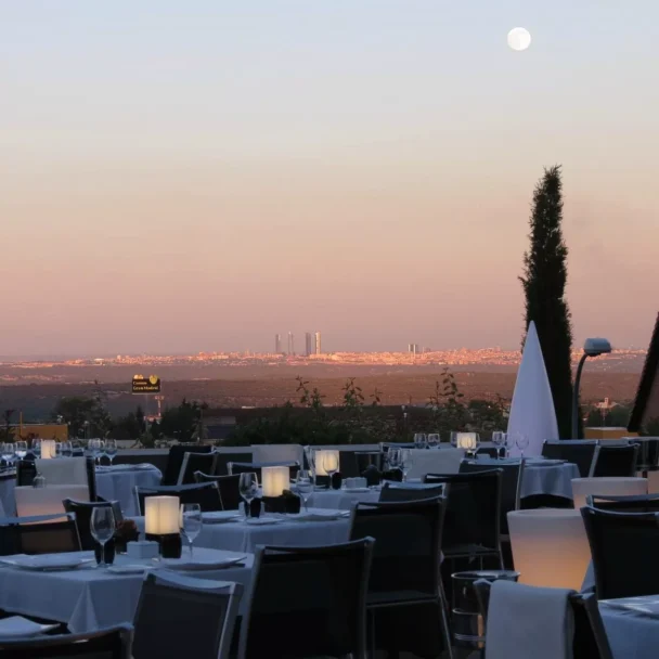 An outdoor restaurant with neatly set tables and chairs overlooks a cityscape under a calming evening sky. A full moon is visible in the sky, casting soft light, and tall buildings can be seen in the distance. Two tall trees frame the view. - Hausion