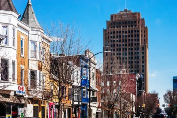 A city street scene with various colorful storefronts and older brick buildings. In the background, a tall modern skyscraper towers over the shorter buildings. Bare trees line the sidewalk under a clear, blue sky. - Hausion