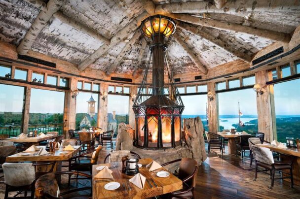 A rustic restaurant interior with a high, wooden ceiling and large panoramic windows offering stunning views of a lake and forest. The space features a central, enclosed fireplace, surrounded by dining tables set with white linens, brown chairs, and wooden floors. - Hausion