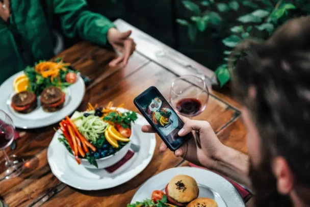 A person photographing a plate of food with a smartphone at a wooden table. The table is set with several colorful dishes, including salads and burgers, and a glass of red wine. Another person's hands are visible in the background, gesturing as they talk. - Hausion