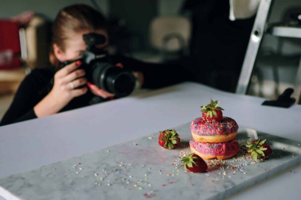 A person is photographing a stack of three colorful, sprinkled donuts topped with strawberries on a marble surface. Several strawberries and sprinkles are scattered around the donuts. In the background, there are blurred objects, adding depth to the image. - Hausion