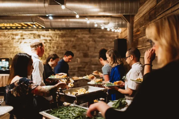 A group of people are standing in line at a buffet in a rustic setting with exposed brick walls and wooden panels. They are serving themselves food from chafing dishes filled with various dishes. String lights hang from the ceiling, creating a warm ambiance. - Hausion