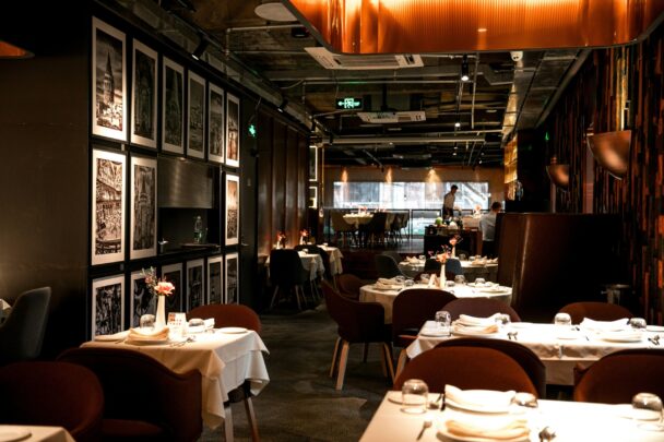 A modern, dimly-lit restaurant with elegant decor and neatly set tables. Several framed black and white pictures adorn the left wall. The dining tables are covered with white tablecloths, and soft lighting creates a warm ambiance. A person is visible in the background. - Hausion