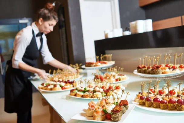 A waitress in a black apron and white shirt arranges several plates of assorted appetizers and hors d'oeuvres on a transparent table. The appetizers include skewers with various ingredients like tomatoes, cheese, and meats, presented on small, decorated picks. - Hausion